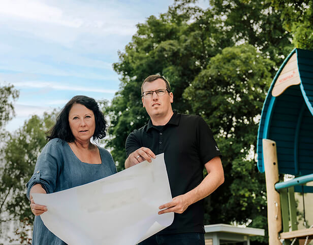 Marion Thiede und Daniel Schietinger stehen mit einer Zeichnung vor einem Spielgerät.